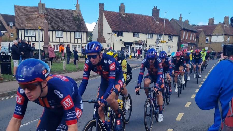 A string of cyclists pass through the centre of Toddington with old buildings behind them and villagers filming from the green using their phone.