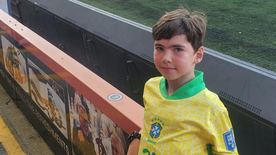 Reuben standing beside a barrier at a sports stadium. He is wearing a yellow and green football jersey with the number 20, resembling the style of the Brazilian national team. The barrier displays advertisements or images related to food and drinks, and the field behind them is covered in turf.