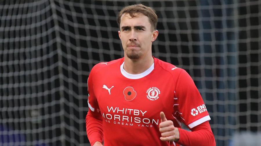 Lewis Billington in action for Crewe Alexandra. He is wearing a red home shirt and is jogging away from goal with the net visible, but blurred, in the background.