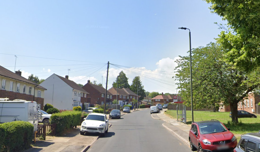 Mickleham Road with houses on the left with cars parked on the pavements lining some of the street and grass on the opposite side to the right with a bus stop in the distance.