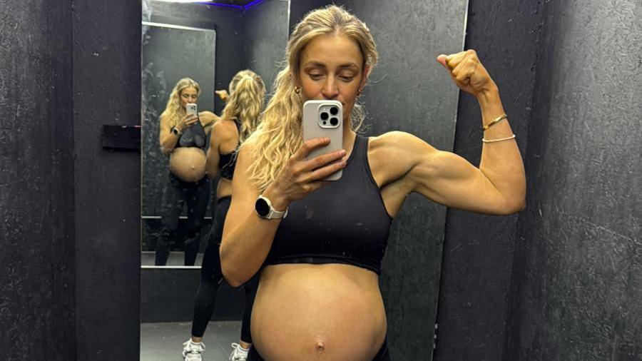Becki Barratt, who is wearing a sports bra so her baby bump is on display, is posing for a selfie in front of a mirror. She has flexed her left arm and holds the camera phone in the other. 