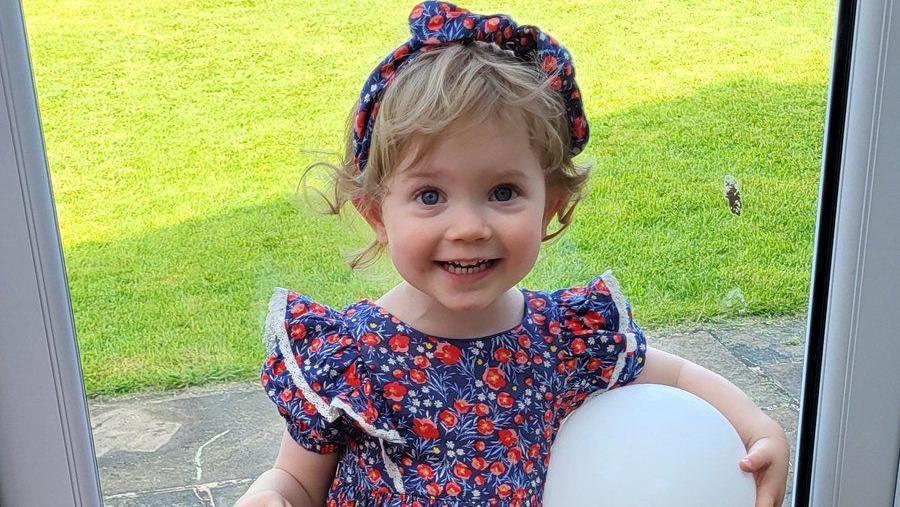 Isabelle Cooper holding a balloon at the door into the garden. She is wearing a blue and red floral dress with matching headband. 