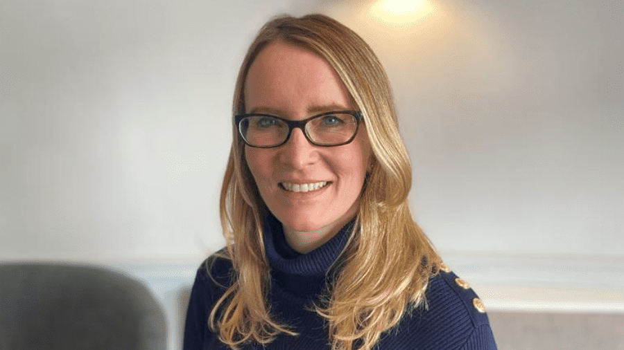 A woman with shoulder-length blonde hair smiling at the camera. She is wearing a navy ribbed polo-neck jumper with gold buttons on the shoulder and black rimmed glasses. She is standing in a room with beige walls.