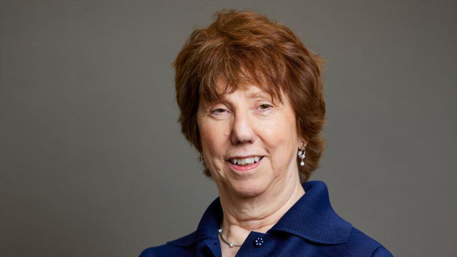 Baroness Catherine Ashton wearing a navy blue shirt smiling at the camera. She has short wavy auburn hair and is wearing droplet earrings 