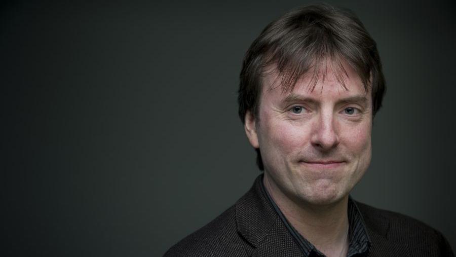 Dr David Clarke is a slim man in his 50s, with straight, dark hair. He is smiling. He is wearing a dark, dog-tooth check jacket and a dark striped shirt. He is standing against a dark grey, lit backdrop.