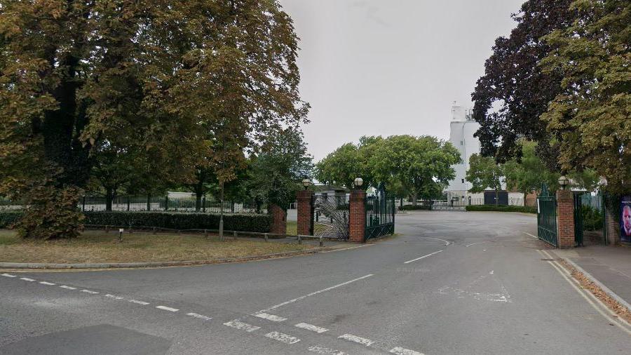 The entrance to Kempton Park racecourse seen in a Google Streetview image. There are large metal gates which are open and one of the stands can just be seen behind some trees.