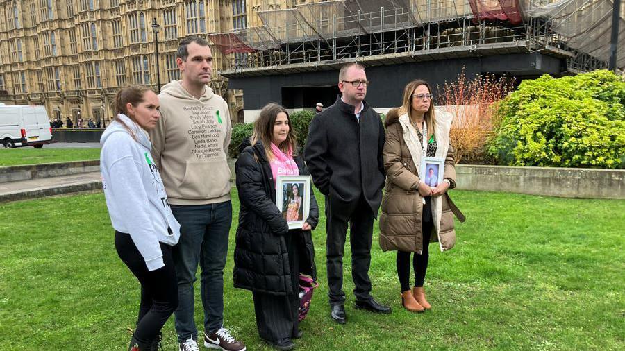 Charlotte and Michael Harnett stand on the green outside the houses of parliament with three other family members. Some are holding up framed photos of their loved ones.