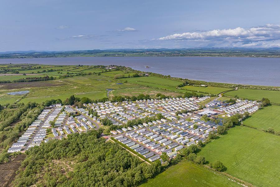 An aerial shot of Glendale Holiday Park. It shows rows of caravans neatly laid out across the site, surrounded by green fields and shrubs, next to a large body of water.