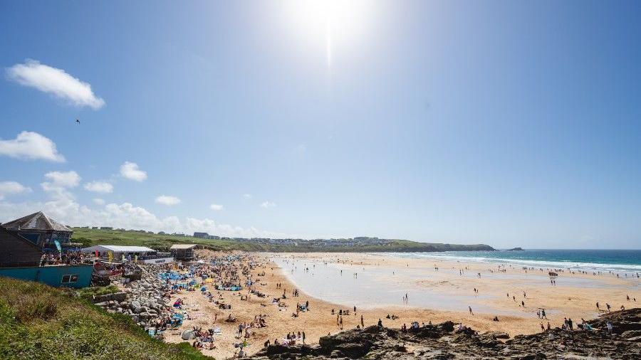 Fistral beach with lots of campers and people enjoying the sun and the sea. The sun is in the sky and there are some clouds but the sky is blue