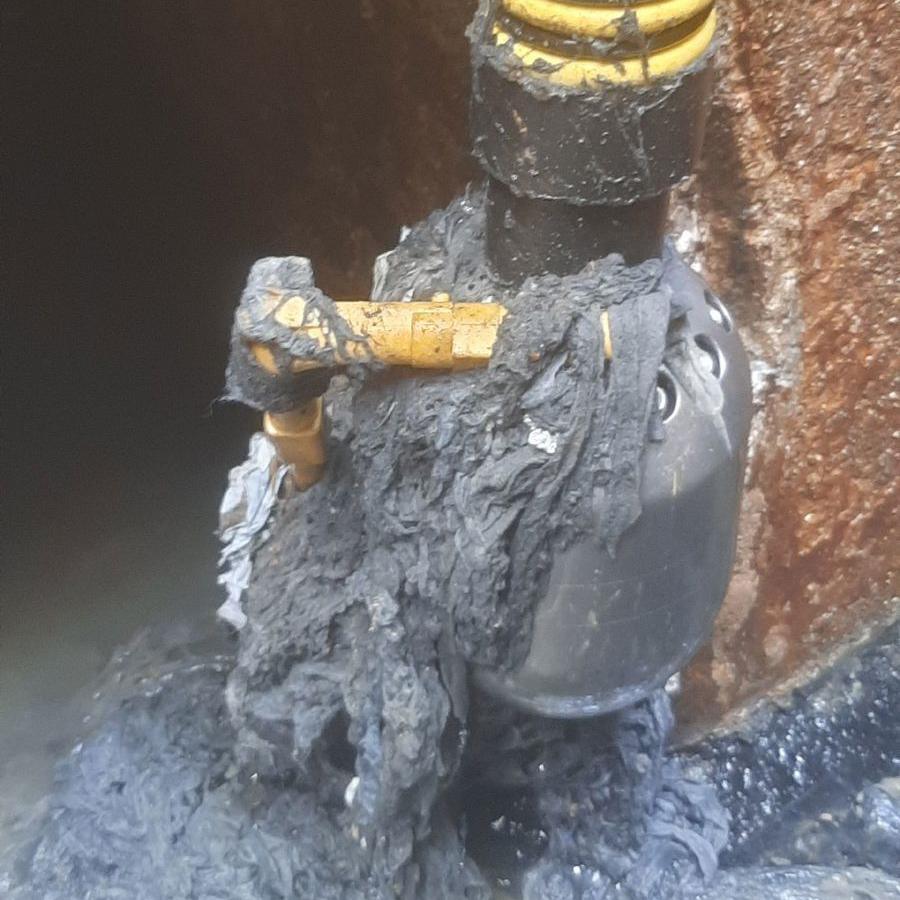 A close-up of a drain pipe valve coated and tangled with congealed wipes and thick black grease.