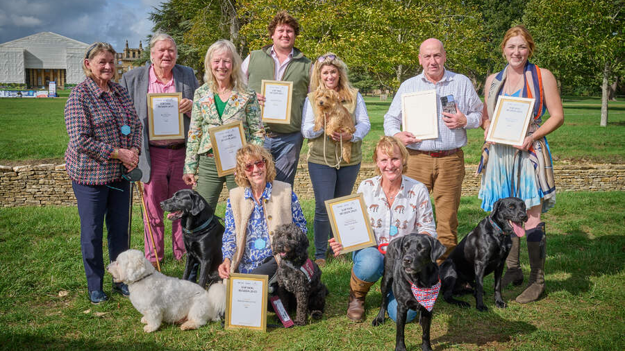 Linda wearing a blue blouse with Gio who is a small black dog with the other award winners stood on grass holding certificates.