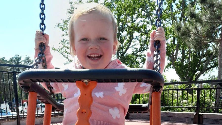 A little toddler on a swing. She has short wispy blonde hair and is beaming at the camera.