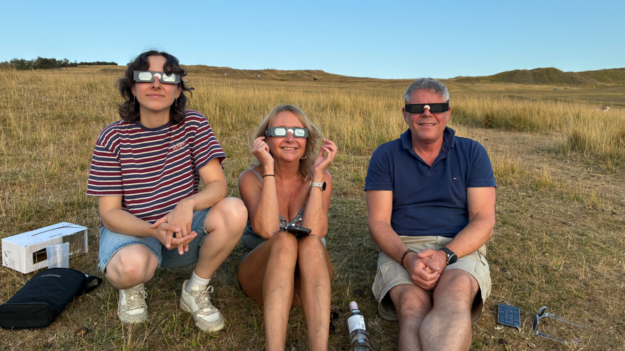 Three people are sat on grass wearing eclipse glasses