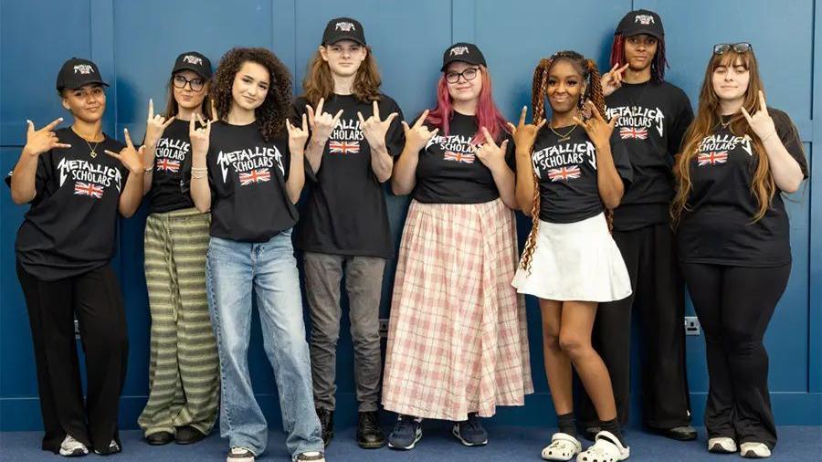 A group of students look to camera and make rock hands whilst wearing t shirts saying Metallica Scholars 