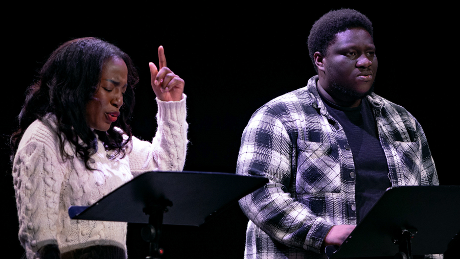 Two people are stood side by side behind black lecterns on a stage. A woman is on the left with medium length black hair, and is wearing a white jumper with one arm in the air. A man on the right is wearing a black T-shirt and tartan jacket.