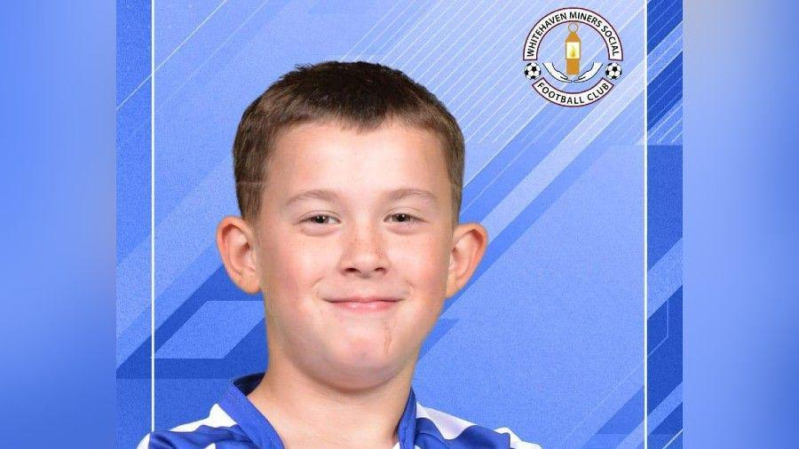 Jay Cartmell, in a blue and white football shirt, looking at the camera and smiling. Behind him is a blue background, and a badge of the Whitehaven Miners Football Club on the top right.