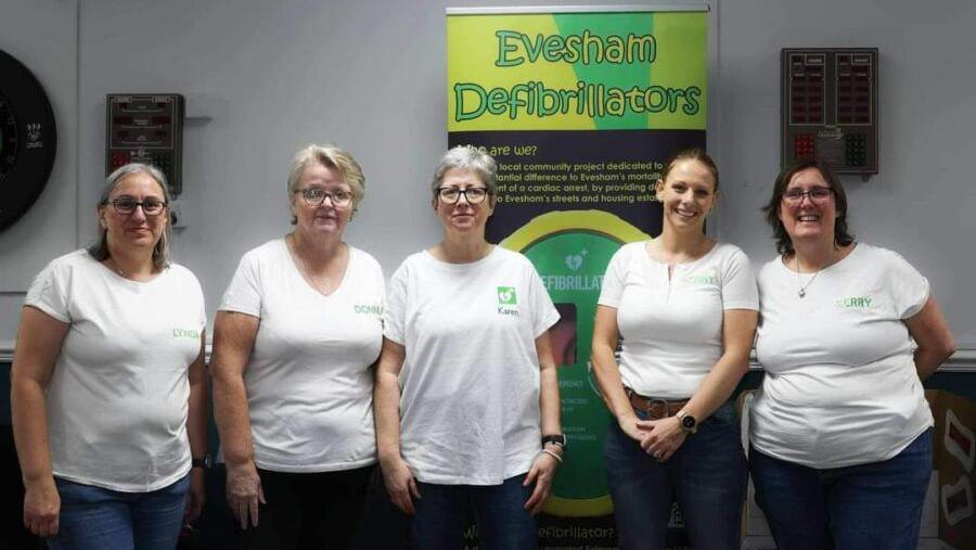 Five women wearing white T-shirts and jeans are standing in front of a sign that reads "Evesham Defibrillators". 