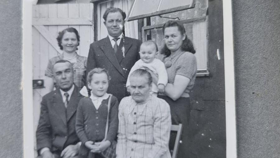 A black and white photo of a family - consisting of two men, three women, a young girl and a baby - smiling outside a Nissen hut.