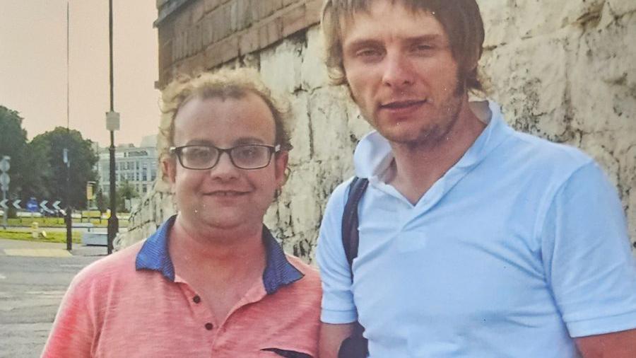 Two men stand side by side. The man on the left has curly light hair, glasses and a pink shirt. The man on the right wears a white polo shirt and a mod haircut.