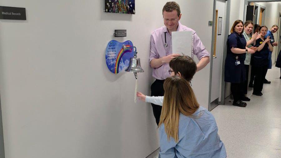 Dulcie has short brown hair and is reaching towards a silver bell that is on a hospital wall. Her mum is crouched next to her and a male hospital worker is standing smiling down at them. In the background, nurses line the corridors and are clapping.