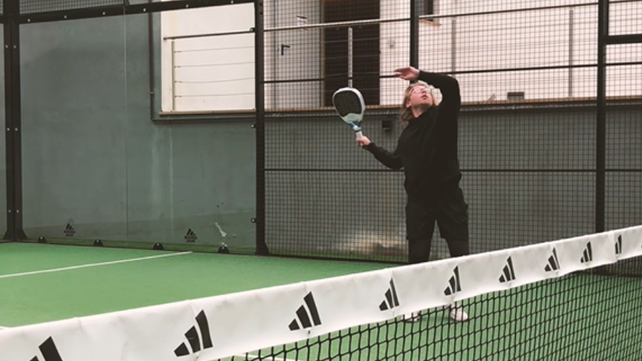 Tom Hutchins in a black sweatshirt and black jogging bottoms, he is positioned holding the padel racket up. He is in an indoor paddle court. The floor below him is green and the wall is grey. 