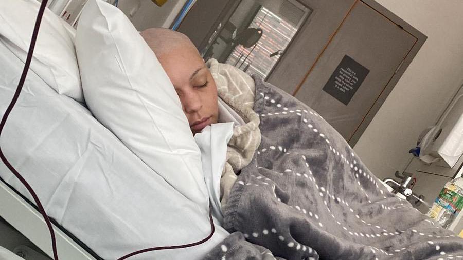 Sacha Gomez, in a hospital bed, in a hospital room, with a brown blanket on her, she is asleep, with a bald head. He is resting on a white pillow.