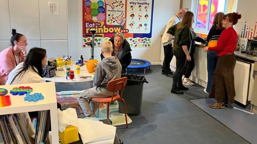 A room with several children sitting at a round table carving pumpkins with adults looking after them.