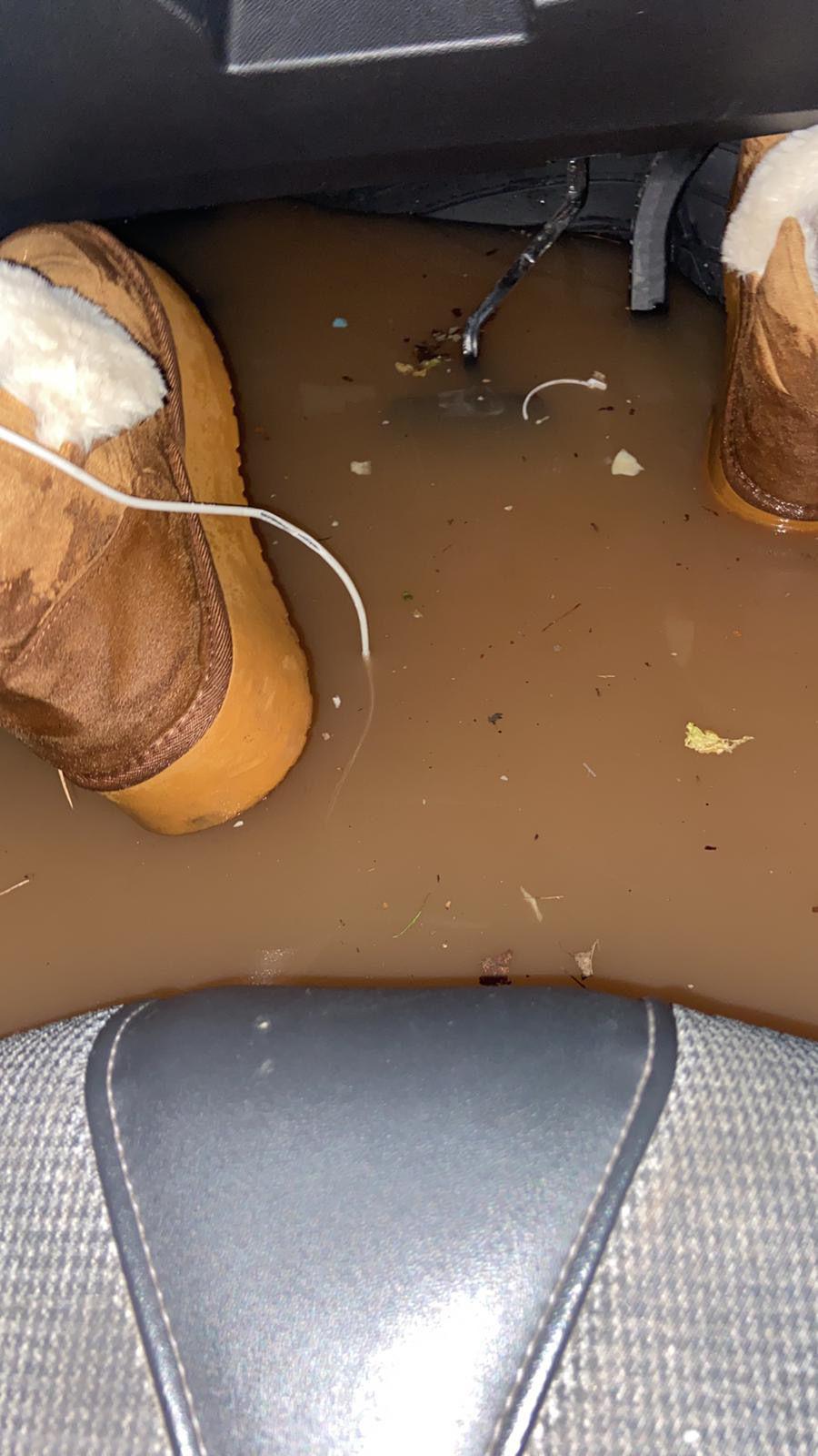 A flooded footwell in a car. A pair of feet in boots are sitting in muddy water in front of the seat.