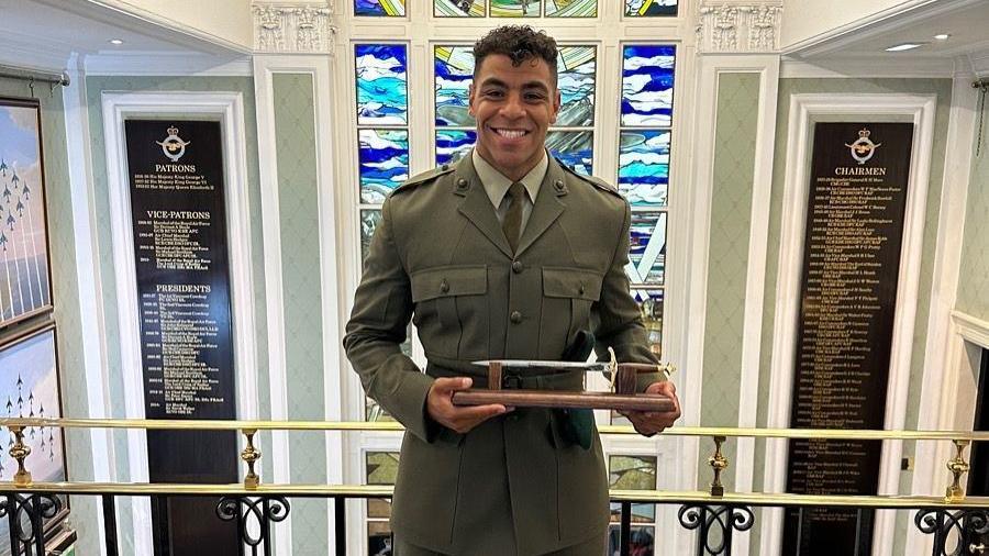 Taylor Lawrence, smiling in a military uniform, stands in front of a stained glass window, with wooden mounted lists of names either side of him holding a small metal trophy-style knife.