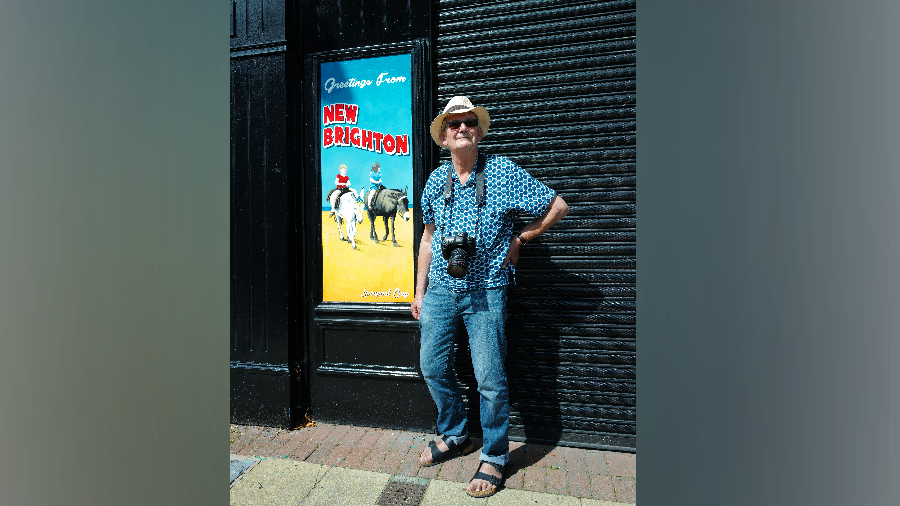 A man wearing a hat and sunglasses, jeans and a summer shirt, with a camera around his neck, stands in the sunshine, next to a building with shutters down and a colourful beach sign which say's New Brighton 