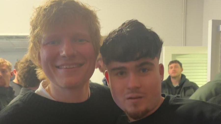 Ed Sheeran poses for a selfie with a young man. They're in a crowded room with other young people.