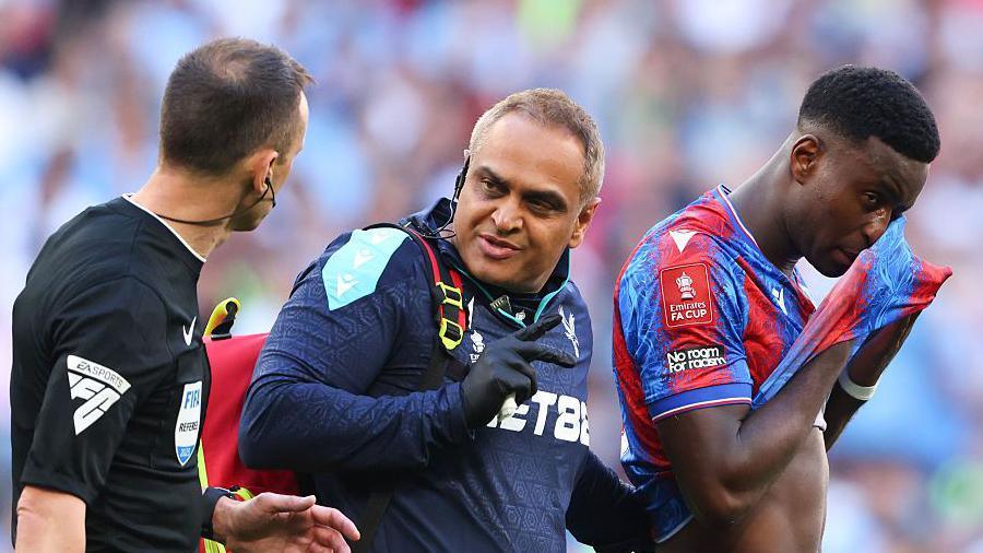 Imitiaz Ahmad talks to referee while helping Marc Guehi off pitch in FA Cup final
