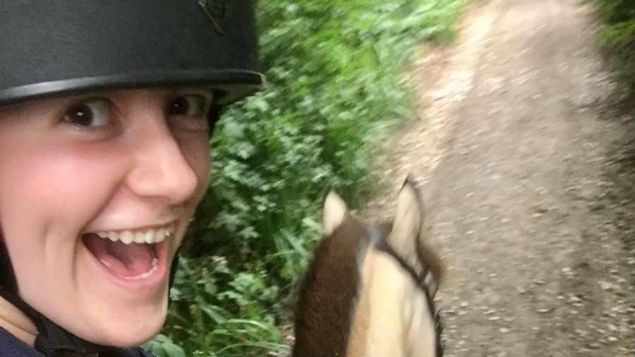 Alys Harrison riding a horse. She is taking a selfie and making a funny face. She has a helmet on. 