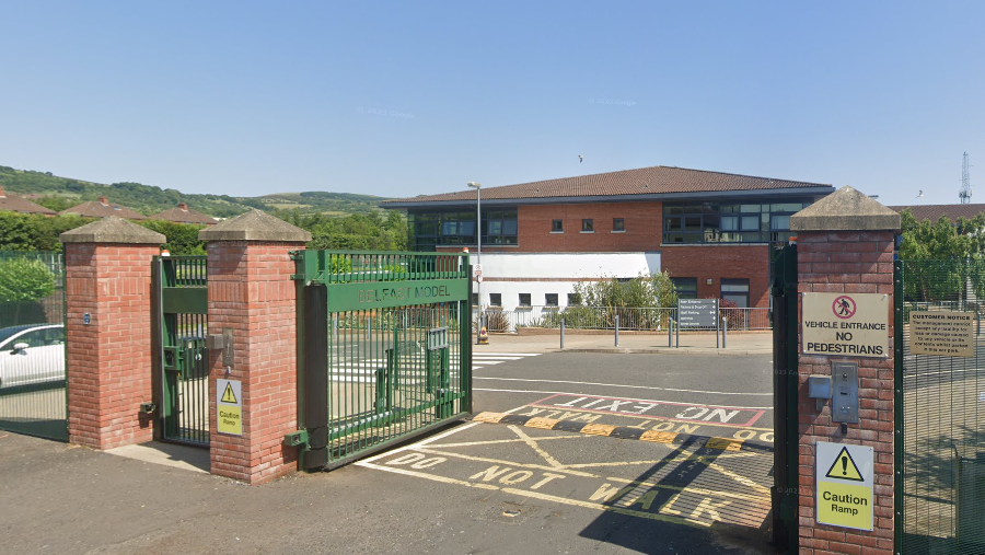 A google image taken from behind the gates of the school, the gate pillars are red brick and the gates and fencing is green. Belfast Model School for Girls has a brown roof with red brick and white walls, and black windows.