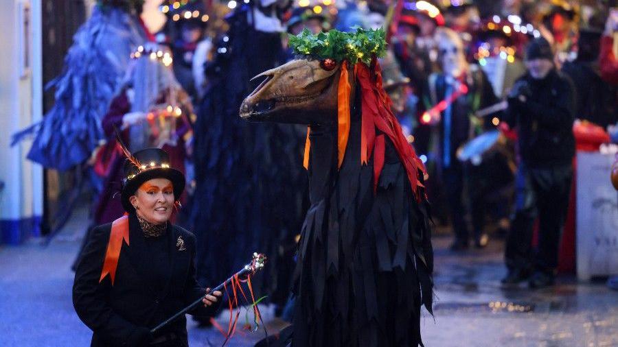 A person seen wearing a hat with lights on, with an orange sash on their shoulder, stood next to someone wearing a full face mask which is extended.