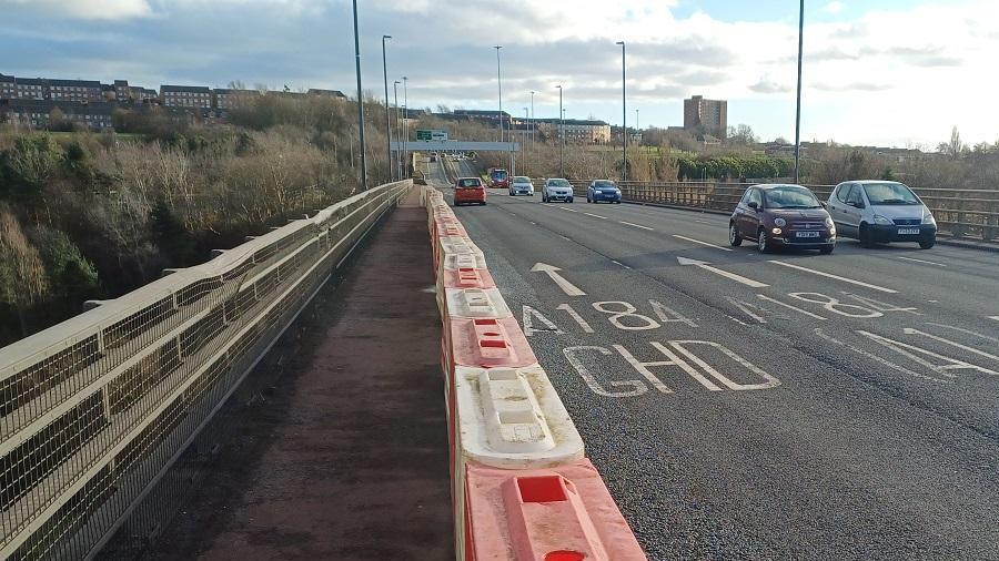 A picture taken from the footpath of the Redheugh Bridge. Cars can be seen travelling on the bridge's four lanes. Red and white plastic safety barriers stand between the footpath and the road.