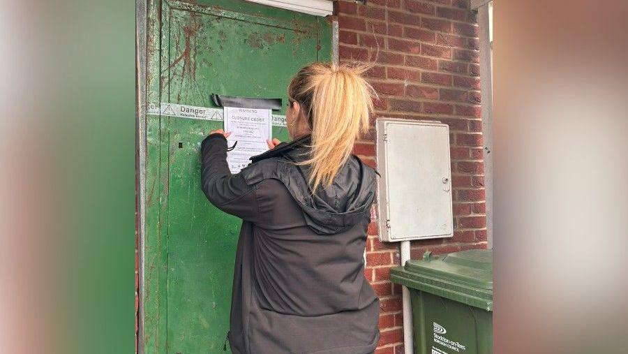 An officer seen from the back, wearing a black jacket, is hanging a closure notice to the door of one of the flats