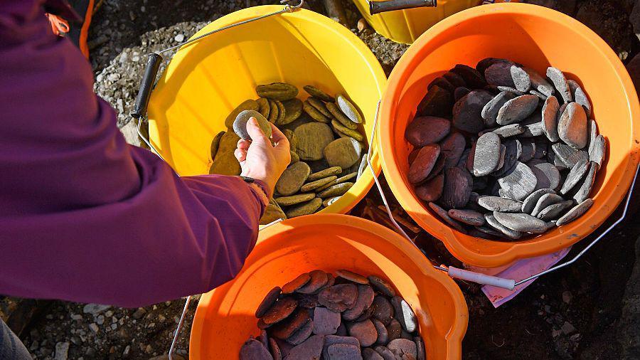 Two orange buckets and a yellow bucket are filled with small flat stones made from slate. Someone in a purple top is picking one up The buckets are on rocky ground.