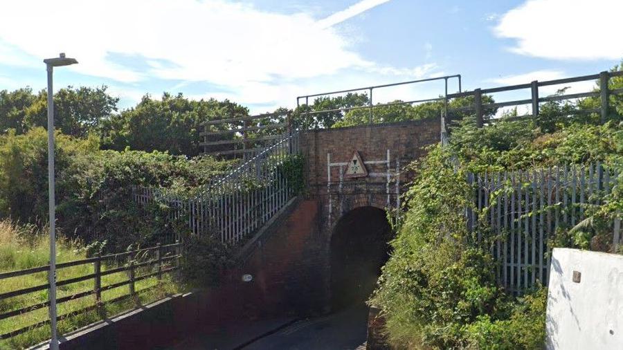 A narrow road passing underneath a small brick railway bridge surrounded by vegetation