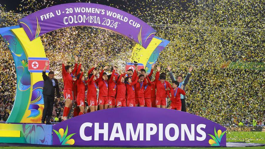 The North Korean under-20s women's squad celebrate on a podium on the pitch that says 'Champions'. They're engulfed in gold-coloured ticker tape.