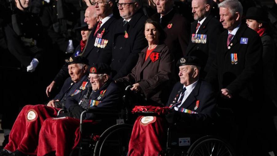 Tre veteranë të Luftës së Dytë Botërore, të veshur me medalje dhe bereta, shtyhen përgjatë Whitehall në karrige me rrota.