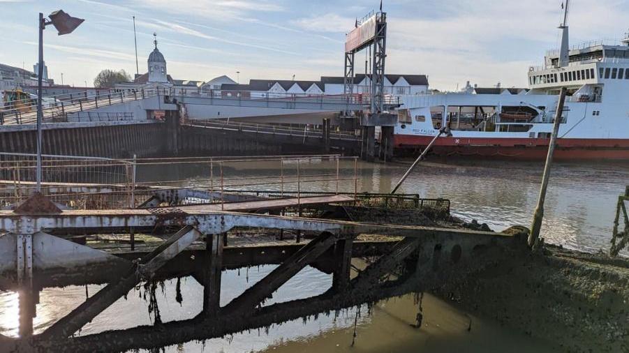 A side view of the Whale roadway section and buffer pontoon of a Mulberry Harbour to the east of Royal Pier at Town Quay in Southampton. It looks in a poor condition. A ferry is seen nearby. It is a sunny day.