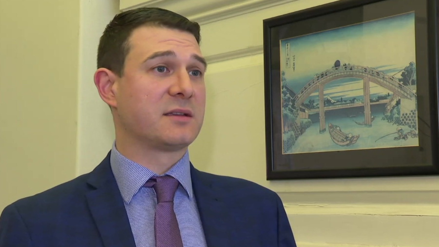 A man in a navy blazer with a lilac shirt and purple tie talks to an off screen interviewer