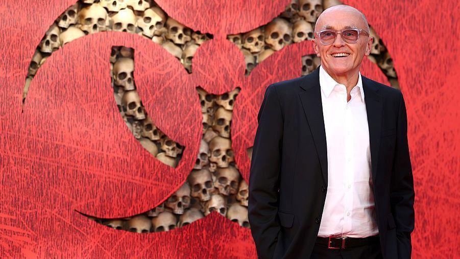 Boyle is wearing a white shirt, black suit jacket and glasses. He is standing in front of a red background with the film's warning sign logo, which features mocked-up human skulls.