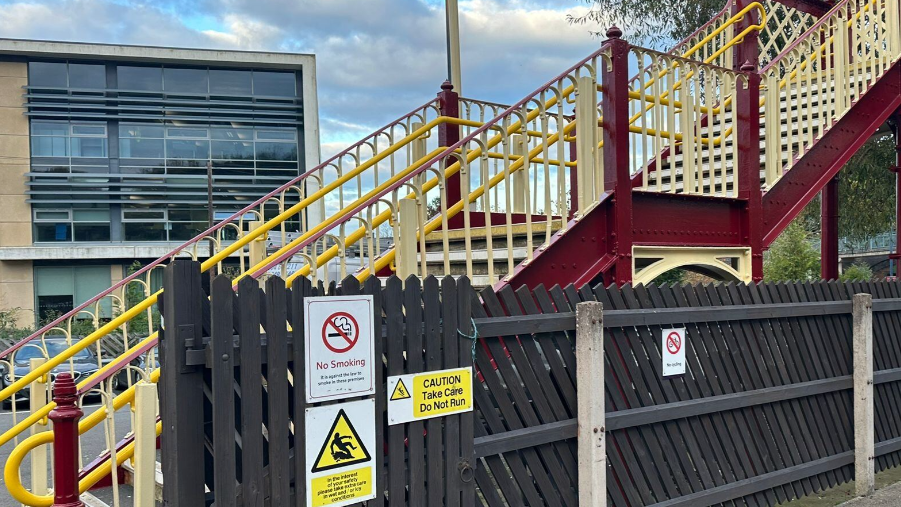 A staircase up to the footbridge behind a black fence with various warning signs attatched