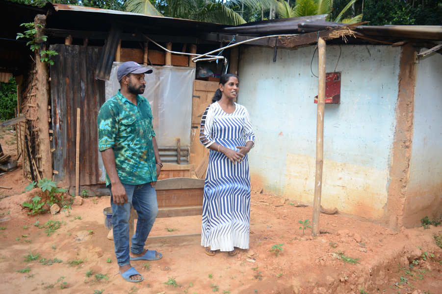 Ravichandran, wearing a cap, green shirt, jeans and blue slip on shoes, stands next to his wife Indrani, wearing a white dress with blue lines, stand outside a building partly made from corrugated iron