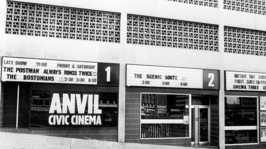 A black and white image of a city street showing three shop units with Anvil Civic Cinema in large letters of one unit's window. All three units have hoardings above the main windows showing film titles and screening times. 