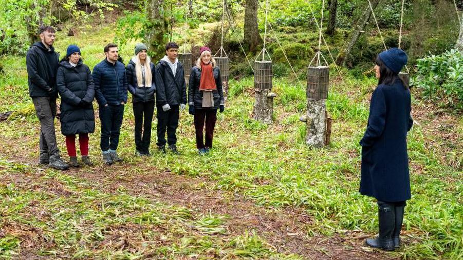 Six contestants from the Traitors stand in a wooded area surrounded by greenery and in front of some hanging baskets. They are all wearing jackets and warm outerwear. Claudia Winkleman is standing in front of them.