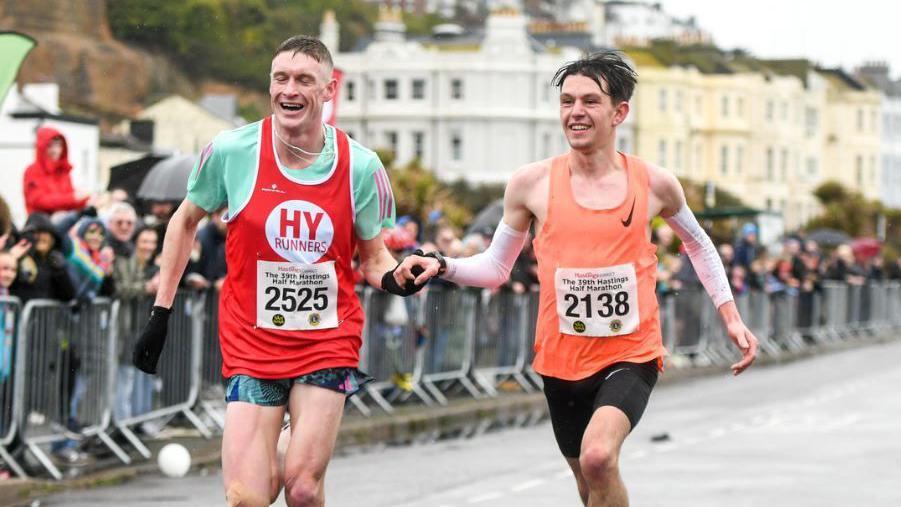 Two male runners in colourful vests and shorts cross the finish line hand in hand looking euphoric