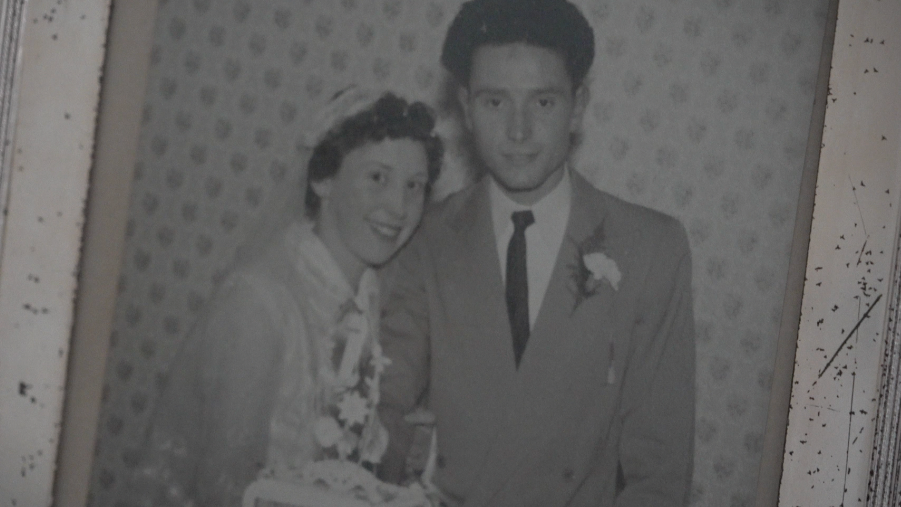 Rose and Harry stand in front of their wedding cake. Rose dressed in a white wedding dress and veil and Harry in a suit with a dark tie on their wedding day 70 years ago.